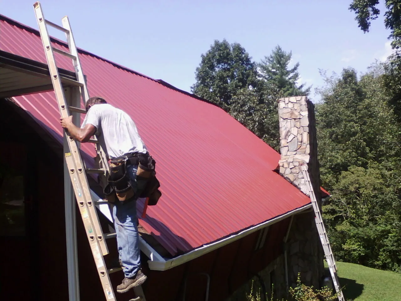 Roofer on a ladder beside a red metal roof and stone chimney — Soffit & Fascia Repair by Roofing Brothers in Elizabethville